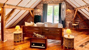 A loft bedroom with a sloped ceiling, exposed brick walls, a bed with pillows, two bedside tables with lamps, and a decorative macrame wall hanging. - Holiday Home 'Am Rande Des Spreewald' with Private Terrace, Private Garden and Wi-Fi (Heinersbrück)