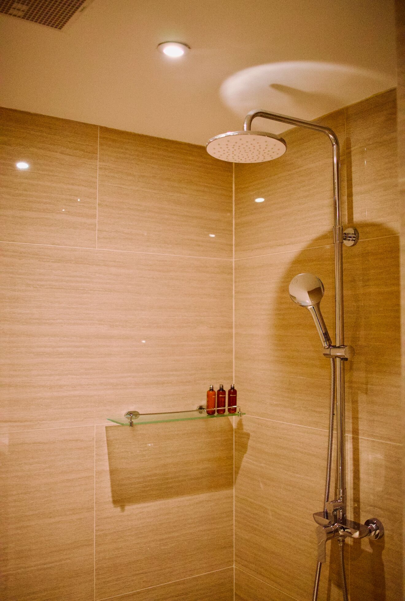 A shower with a handheld showerhead and a wall-mounted rain showerhead. There is a shelf with toiletries.