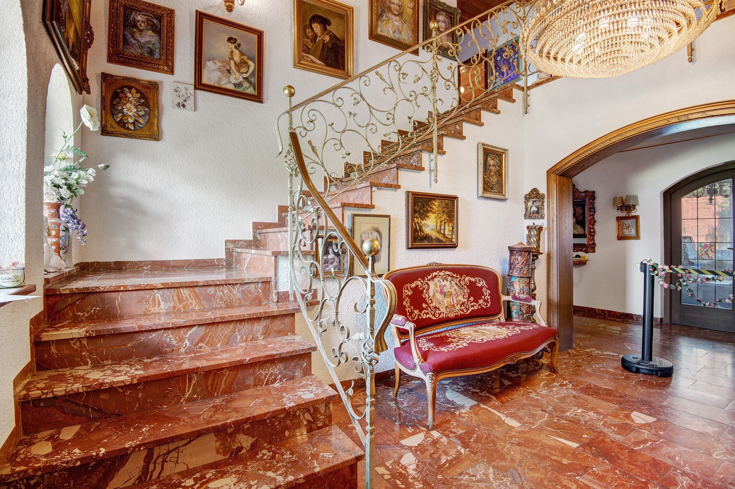 Eine prächtige Treppe mit aufwendiger Metallreling, ein rot gepolsterte Couch und eine Kristallleuchter.