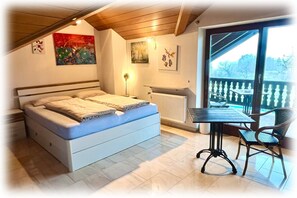 Una camera da letto con un letto, un tavolo con sedie, un termosifone e un balcone con ringhiera.
