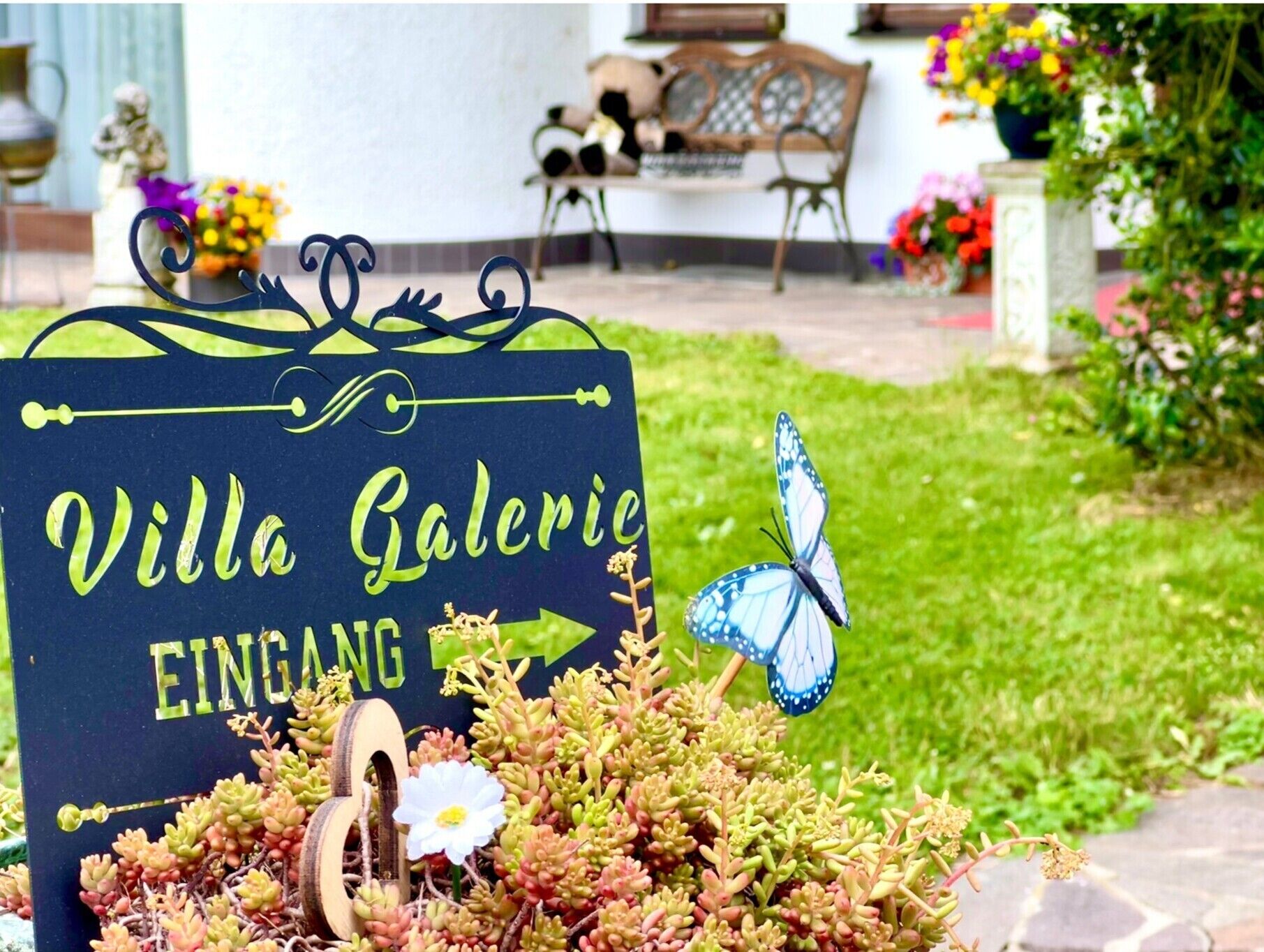 Ein Schild mit der Aufschrift 'Villa Galerie EINGANG', verziert mit einem Schmetterling und Sukkulenten.