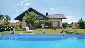 En swimmingpool med solstolar, ett hus med ett tak täckt av takpannor och en gräsklädd trädgård.
