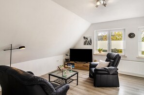 A modern living room with a flat-screen TV, leather sofas, a glass coffee table, and a bookshelf.