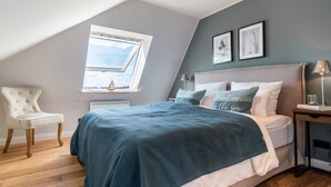 A bedroom with a sloped ceiling, a skylight, a bed with a blue bedspread, a bedside table with a lamp, and a chair.
