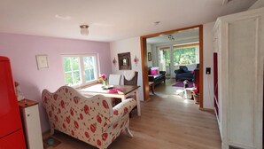 A kitchen with a red refrigerator, a floral sofa, a dining table with chairs, and a view into the living room through an open doorway.