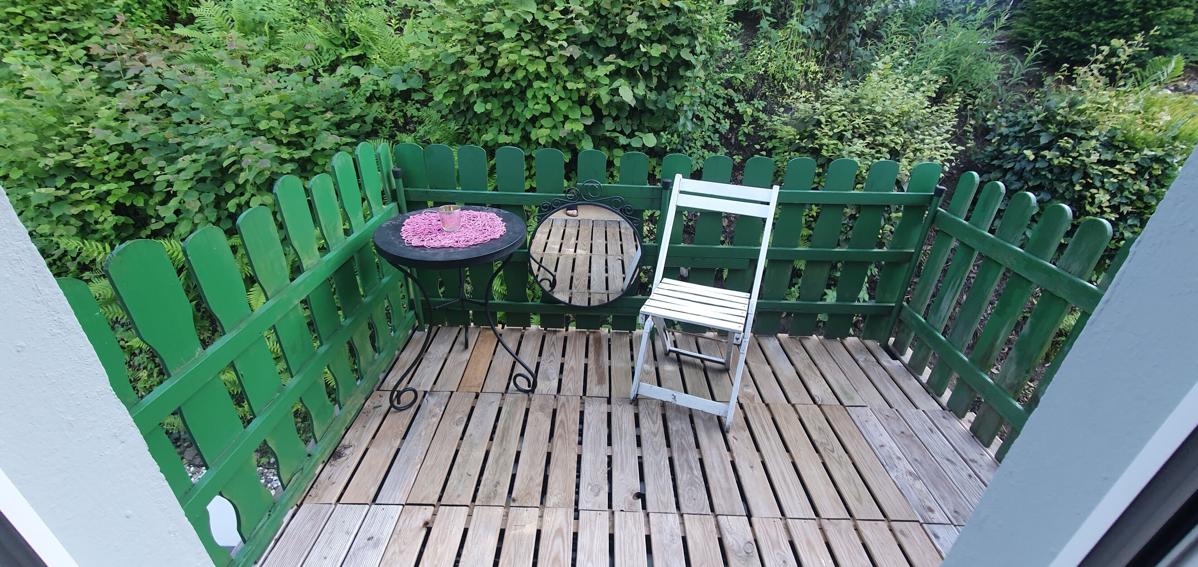 Une terrasse en bois avec une clôture verte, une petite table ronde recouverte d’une nappe rose et une chaise blanche.