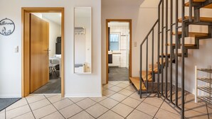 A hallway with a wooden door, a mirror, a staircase with metal railings, and a kitchen in the background. - Apartment 'Celle-Bostel' with Private Terrace, Garden, and Wi-Fi (Celle)