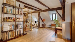A room with a wooden bookshelf, a desk with a computer, a chair, a lamp, and a large window.