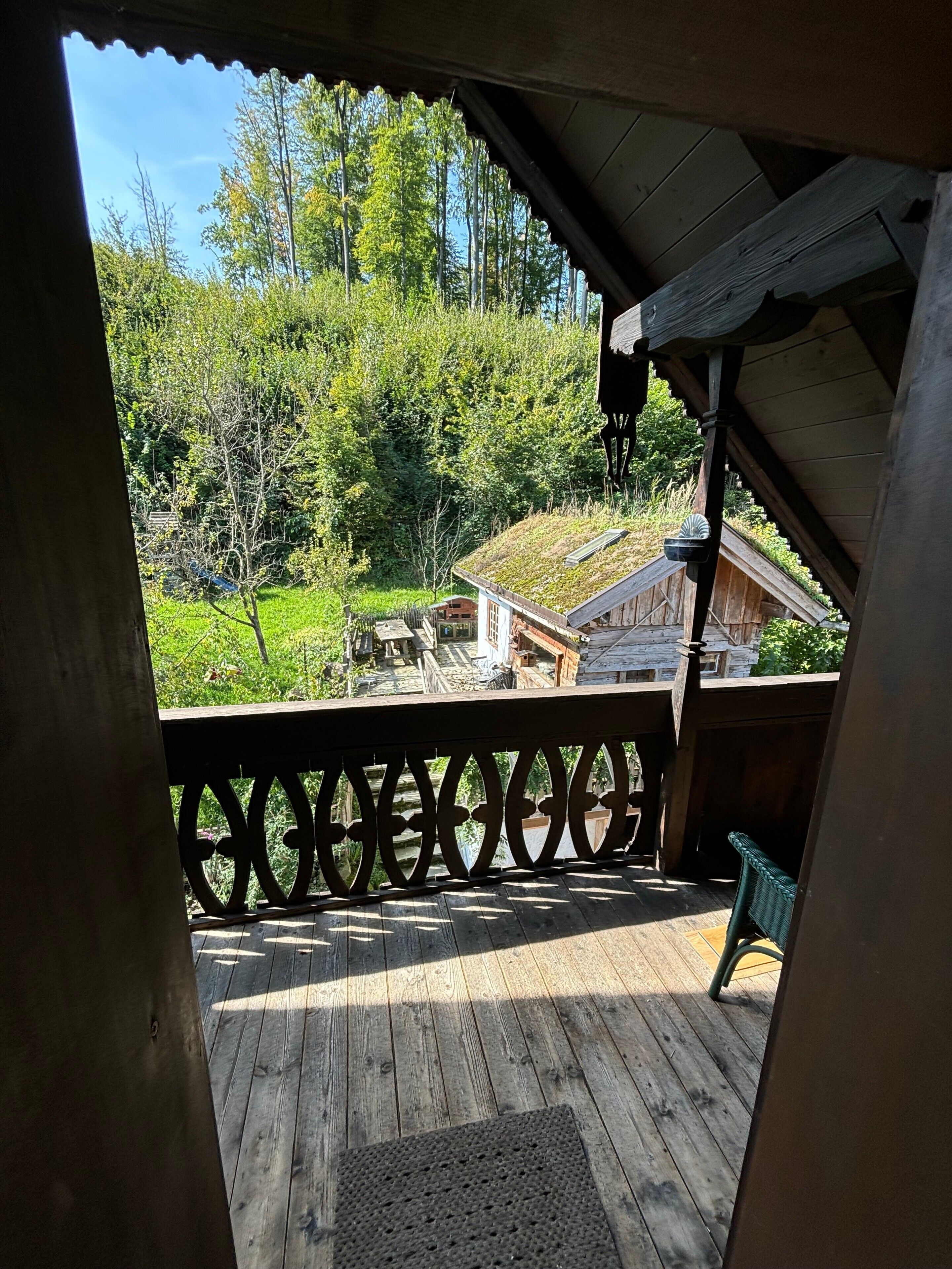 Um deck de madeira com guarda-corpo, área coberta e vista para uma casa com telhado verde e árvores ao fundo.