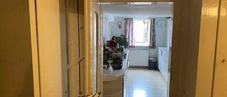 A narrow hallway leading to a kitchen with a radiator, a trash bin, and a window with curtains.