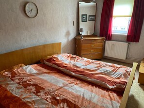Quarto com cama, penteadeira de madeira, relógio na parede, espelho e janela com cortinas.