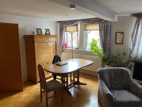 A dining area with a wooden table, chairs, a cabinet, a radiator, a window with curtains, and a plant.