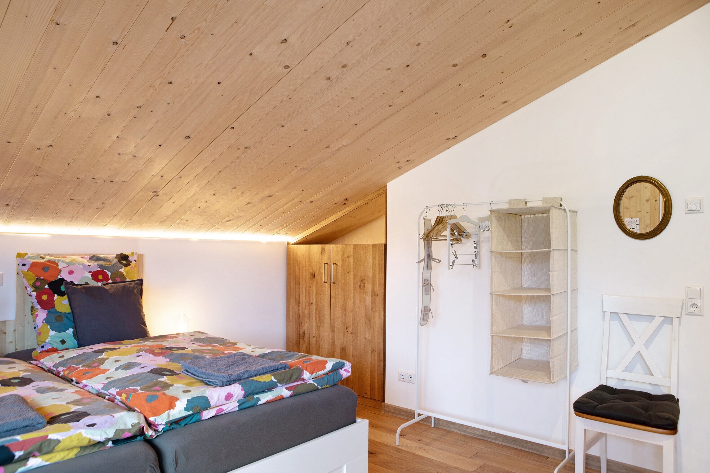 Une chambre avec un plafond en bois, un lit recouvert d’une courtepointe colorée, une armoire en bois, une chaise blanche et une étagère fixée au mur.