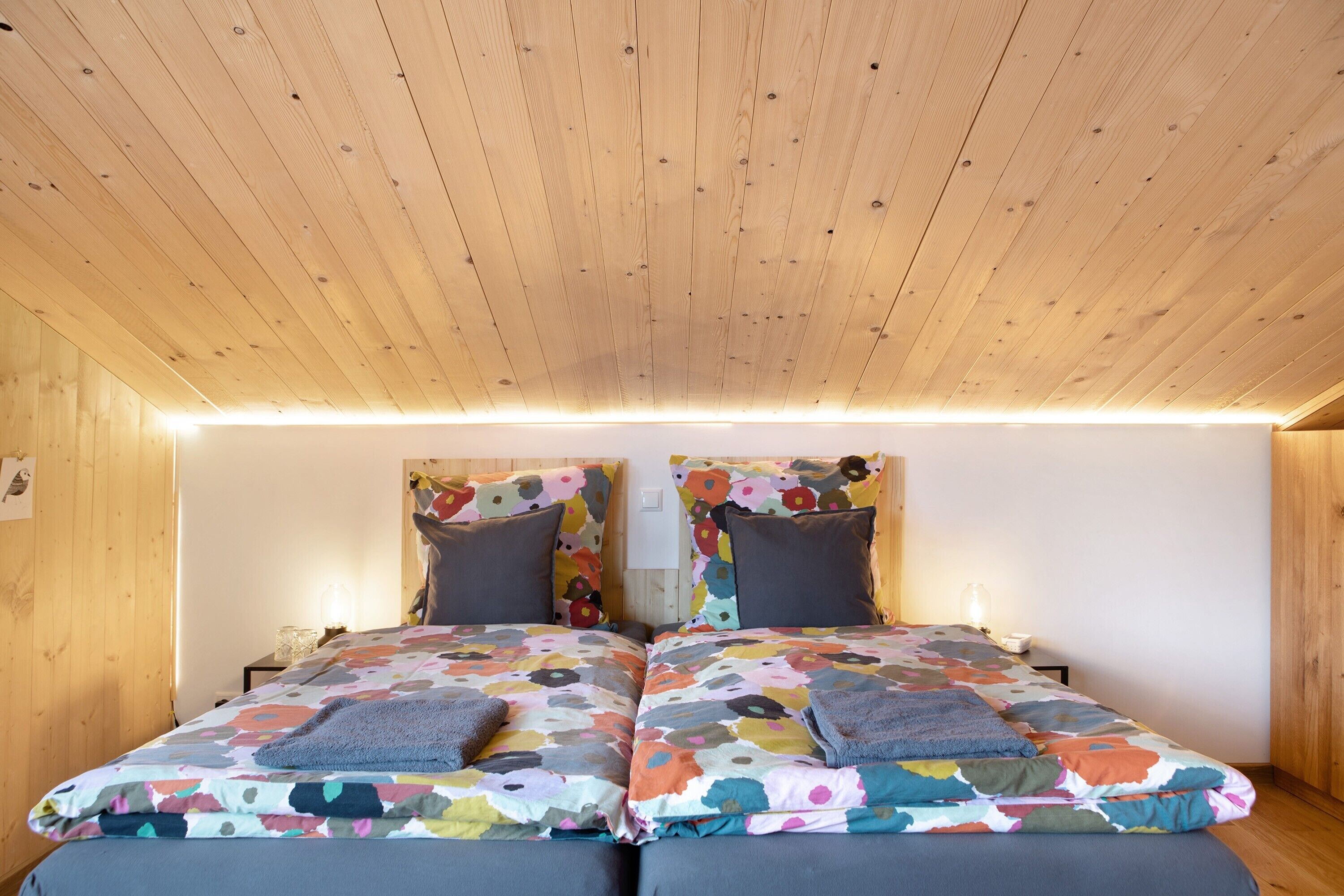 A room with wooden ceiling and walls, two beds with colorful floral bedding, and bedside lamps.