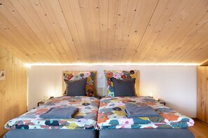 A room with wooden ceiling and walls, two beds with colorful floral bedding, and bedside lamps.
