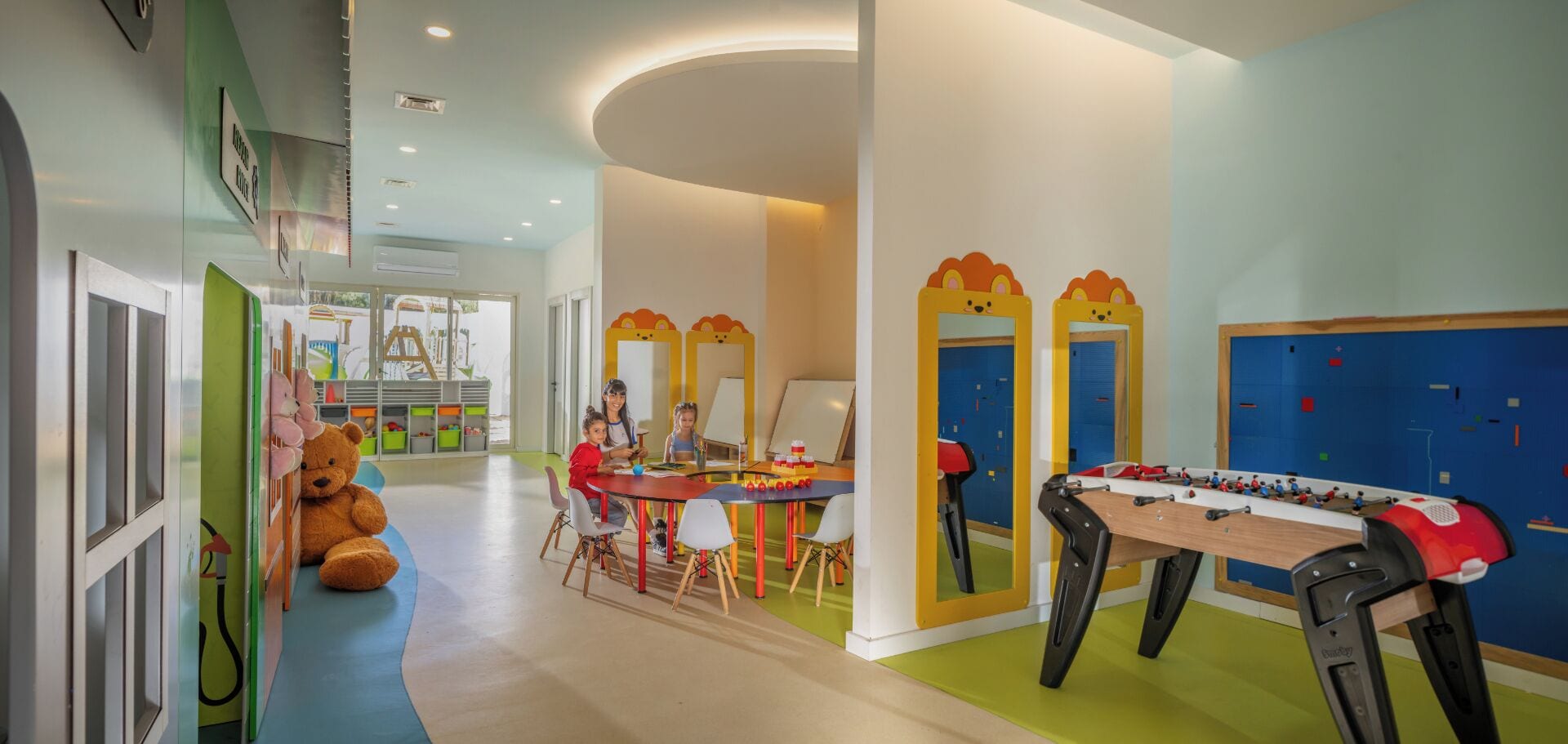 Zona de juegos para niños con mesa de fútbol de mesa, un gran osito de peluche y una mesa con material de arte.