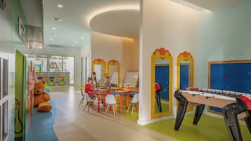 Zona de juegos para niños con mesa de fútbol de mesa, un gran osito de peluche y una mesa con material de arte.