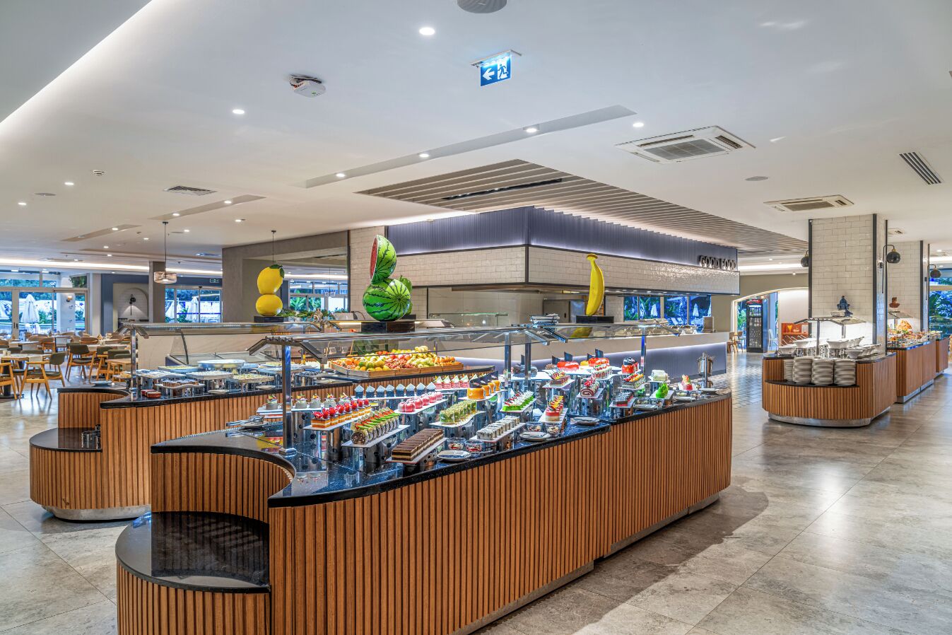 A modern buffet area with a variety of food options, including fruits and desserts, in a spacious and well-lit setting.