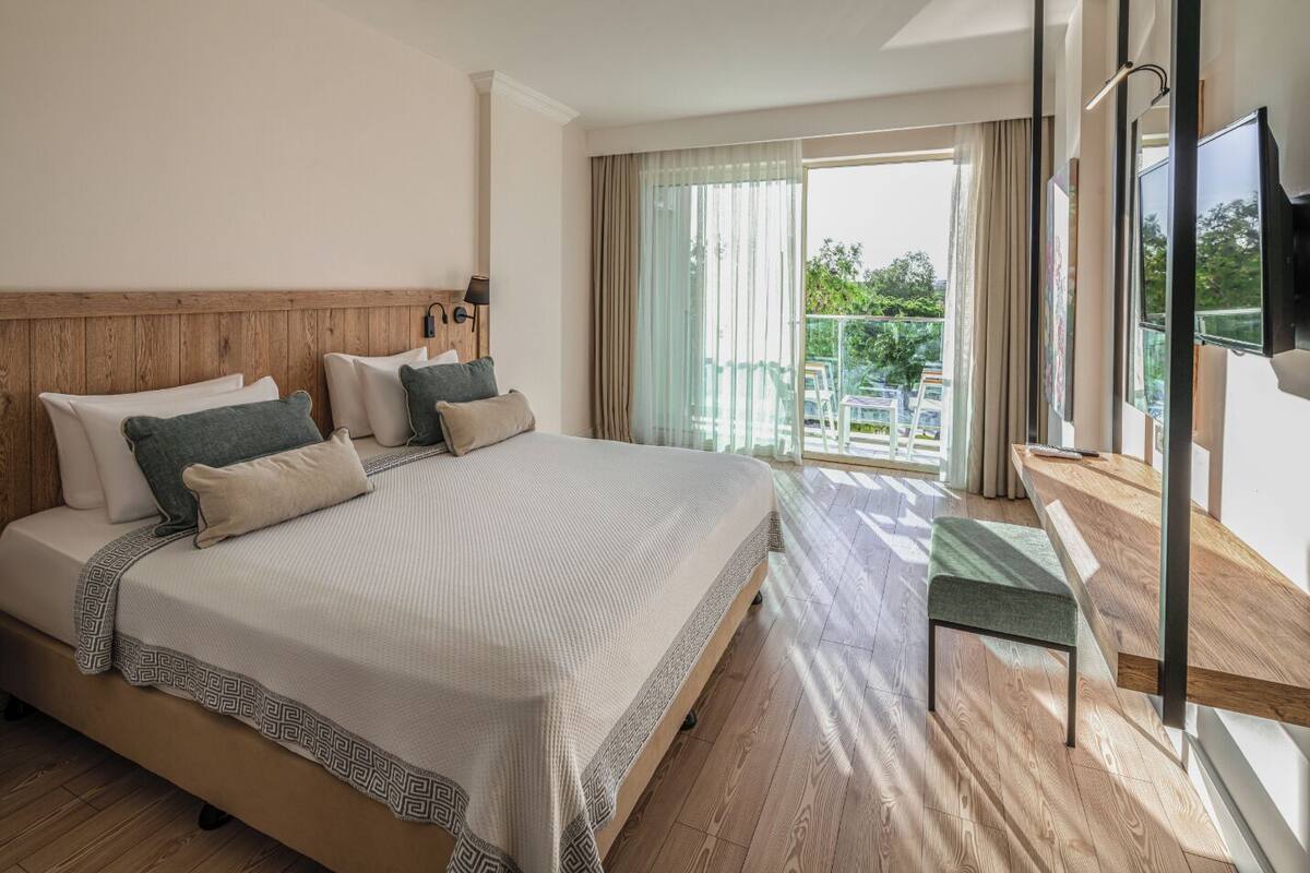 A hotel room with a large bed, wooden headboard, and a balcony with a view of greenery.