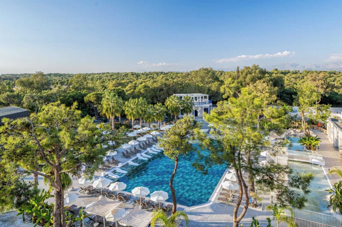 Een resort met een groot zwembad, omringd door ligbedden en parasols, met op de achtergrond een dichte boombeplanting en een heldere lucht.