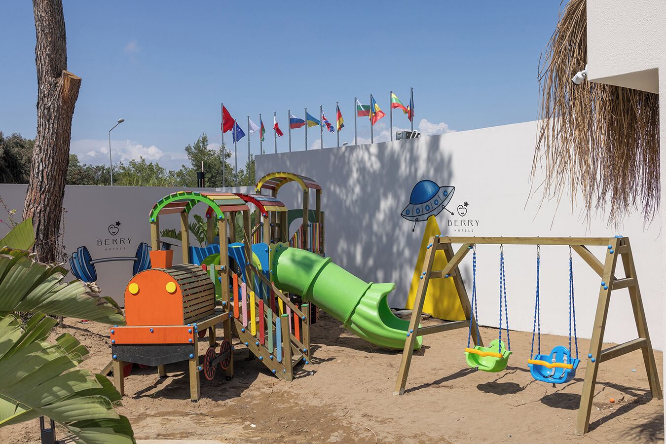 A children's play area with a slide and swings, surrounded by a white wall with flags.