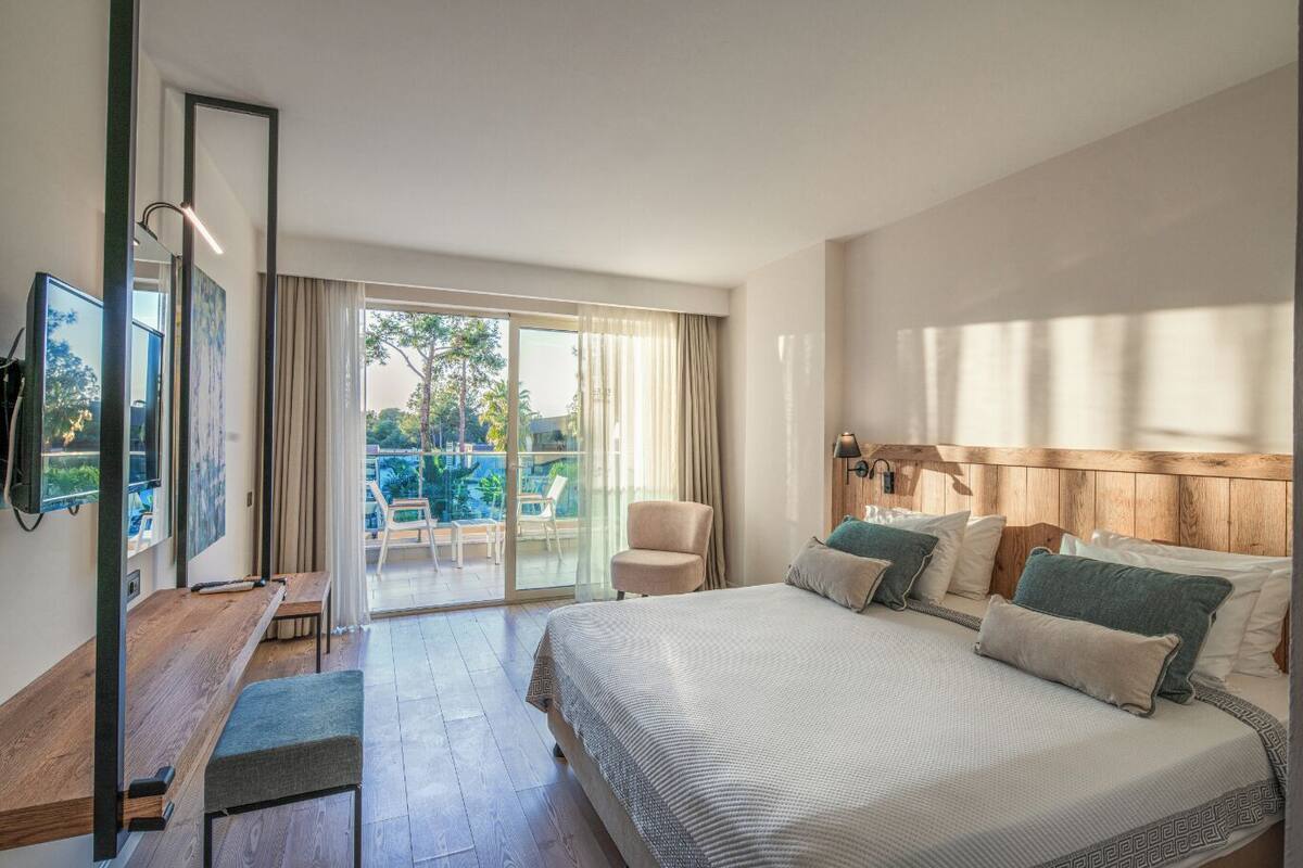A modern hotel room with a large bed, a desk, a chair, and a balcony with outdoor seating.