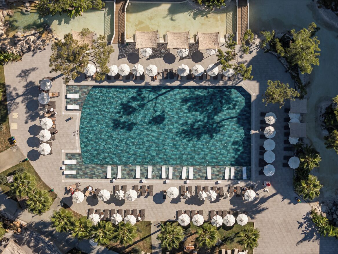 Eine Vogelperspektive zeigt einen Ferienort mit einem großen rechteckigen Swimmingpool, umgeben von Liegebereichen inklusive Sonnenschirmen und Liegestühlen.