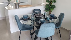 A modern kitchen with white cabinets, a built-in oven, and a sink. There is a dining area with a glass table, blue chairs, and a potted plant. - Penthouses Blue Lagoon (VC)