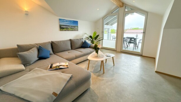 Ein modernes Wohnzimmer mit einem grauen Sofa, einem kleinen runden Tisch und einem großen Fenster mit Blick ins Freie.