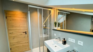 Un baño moderno con cabina de ducha de vidrio, puerta de madera y lavamanos blanco con espejo sobre él.