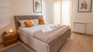 A neatly made bed with white and beige bedding, a wooden nightstand with a lamp, and a radiator on the wall.