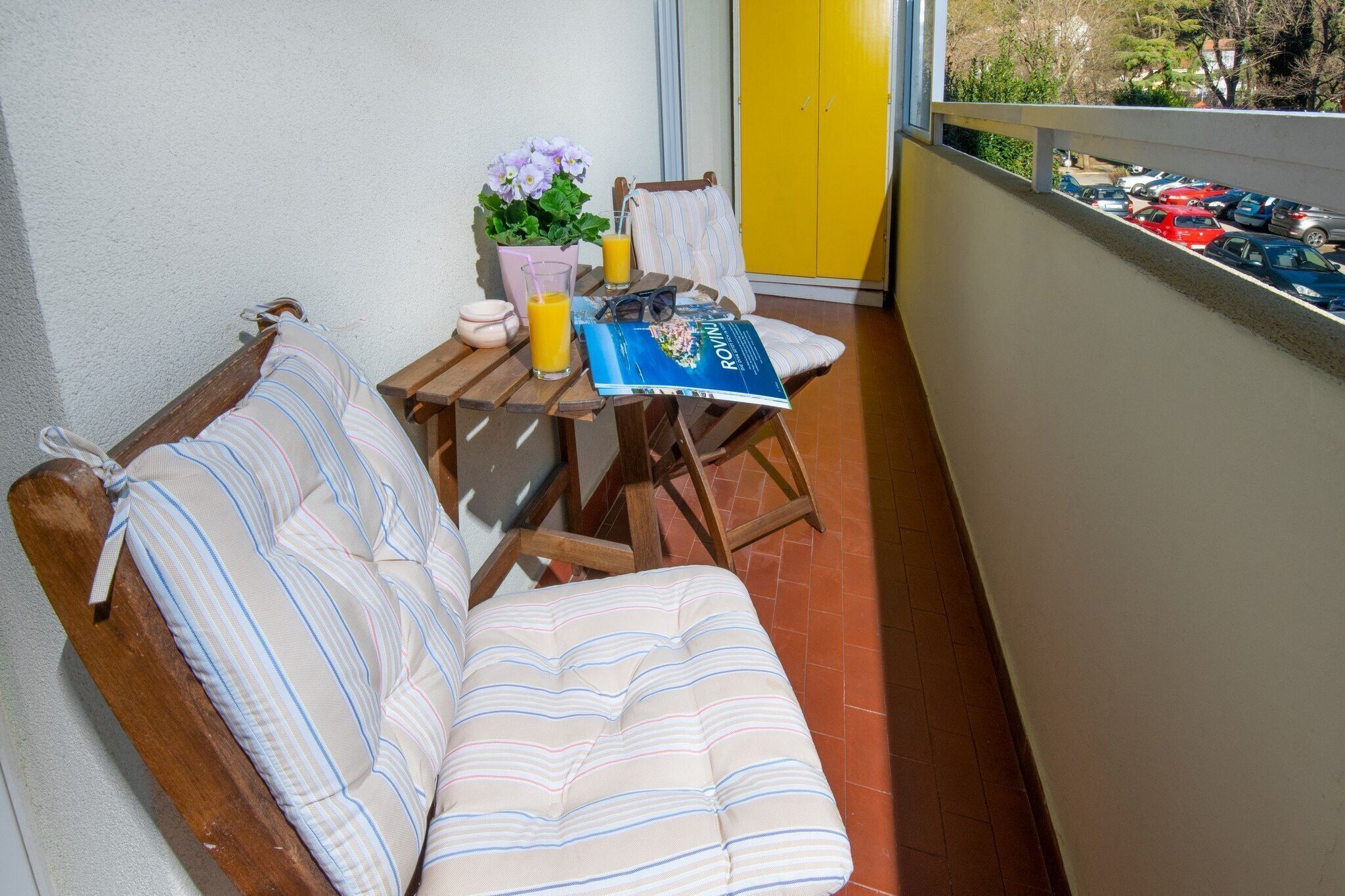 A balcony with a wooden bench, a table with a book, a glass of orange juice, and a potted plant.