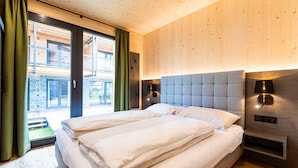 A double bed with white linens in a room with wooden panel walls, a large window with curtains, and two wall-mounted lamps.