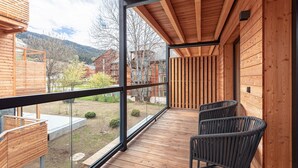Un balcón de madera con barandilla de vidrio, dos sillas de mimbre y vista a un paisaje montañoso.