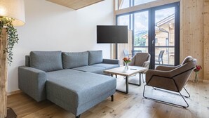 Un salon moderne avec un canapé d’angle gris, une table basse et un fauteuil. Un téléviseur est fixé au mur et une fenêtre offre une vue sur l’extérieur.