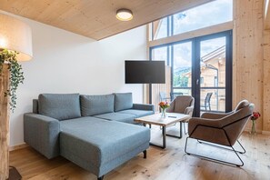 Un salon moderne avec un canapé d’angle gris, une table basse en bois et un fauteuil marron. Un téléviseur à écran plat est fixé au mur, et une grande fenêtre offre une vue sur l’extérieur.