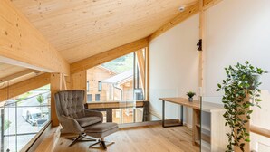 Ein modernes Interieur mit Holzbalkendecke, einem gepolsterten Sessel, einem Glastisch und Blick auf ein Gebäude außerhalb.