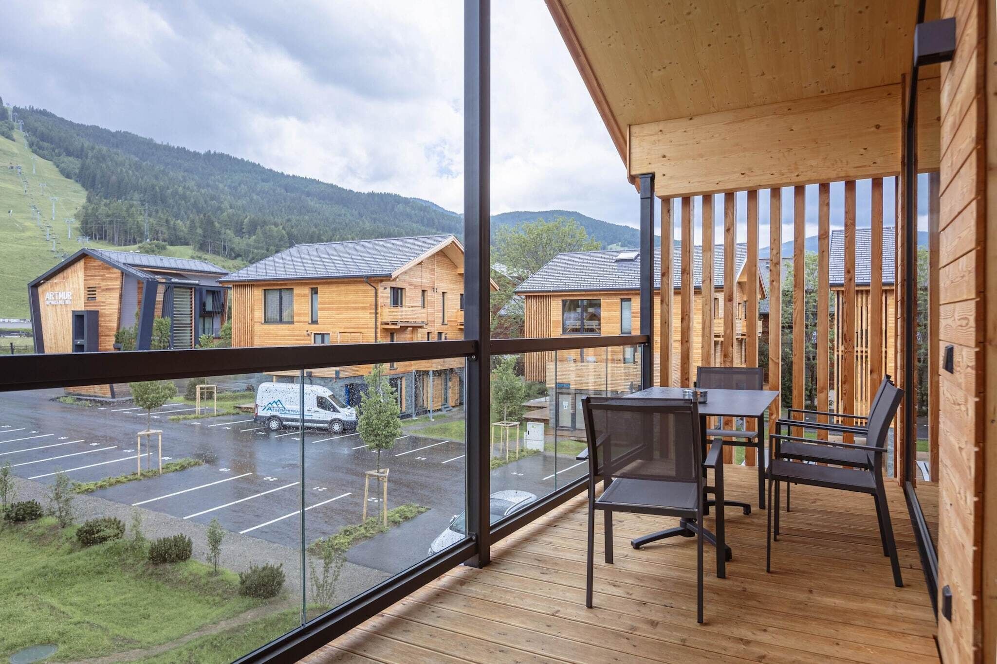 Um deck de madeira com mesa e cadeiras, com vista para um estacionamento e edifícios.
