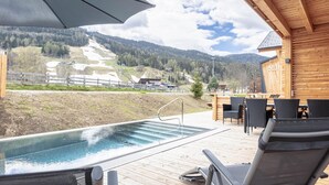 Una terraza de madera con hidromasaje, mobiliario exterior y vistas a un paisaje montañoso.