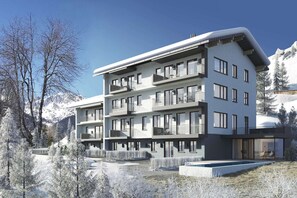 Un immeuble à plusieurs étages doté de balcons, de grandes fenêtres et d’un espace piscine, entouré d’arbres et de montagnes couverts de neige.
