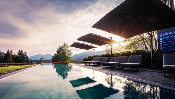 Un área junto a la piscina con sillones y sombrillas, rodeada de árboles y montañas.