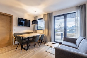 A modern living room with a wooden dining table, grey sofa, and a large window offering a mountain view.