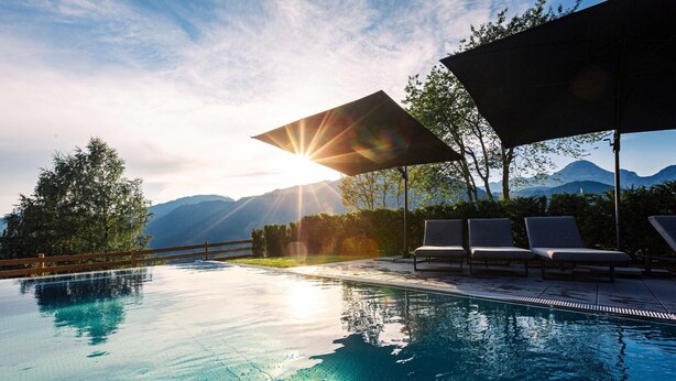 Una piscina infinita con vistas a montañas, sol y una zona de estar cubierta con sillones de descanso.