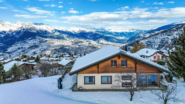 Eine Holzhütte mit schneebedecktem Dach, vor der sich verschneite Berge und ein klarer, blauer Himmel erheben.
