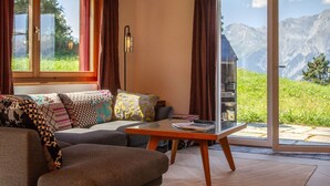 Ein gemütliches Wohnzimmer mit einer Couch, einem Glastisch und Blick auf Berge durch eine Schiebetür.