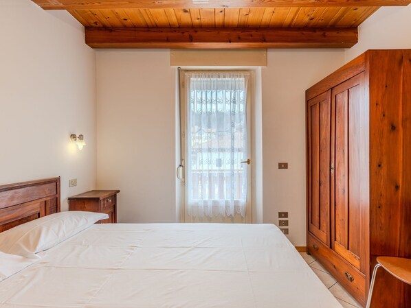 A bedroom with a wooden ceiling, a large wardrobe, a bedside table, and a window with a lace curtain.