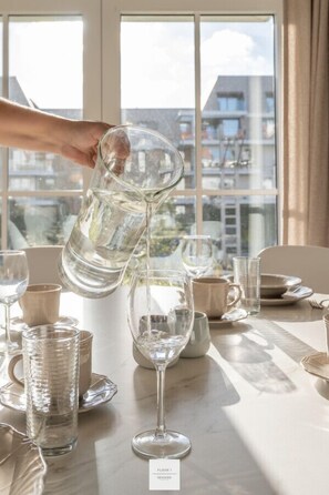A hand pouring water into a glass pitcher on a table with various glassware and a sign indicating a room number and side. - Boathouse Floor, Hamptons style at the harbor (Nieuwpoort)