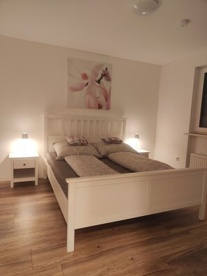 Un dormitorio con una cama blanca, dos mesitas de noche con lámparas, un cuadro grande con motivos florales en la pared y una ventana con vistas.
