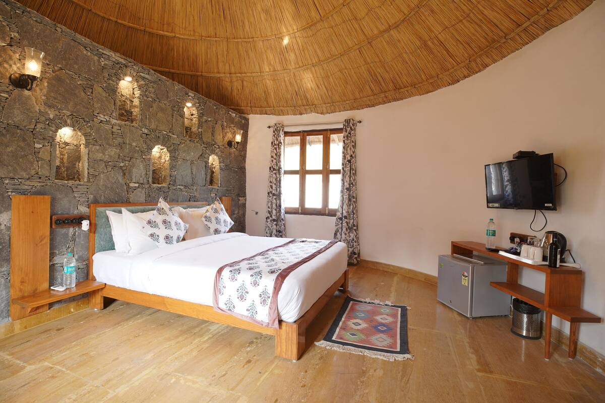 Een slaapkamer met een stenen muur, een houten plafond, een bed, een televisie, een klein bureau en een raam met gordijnen.