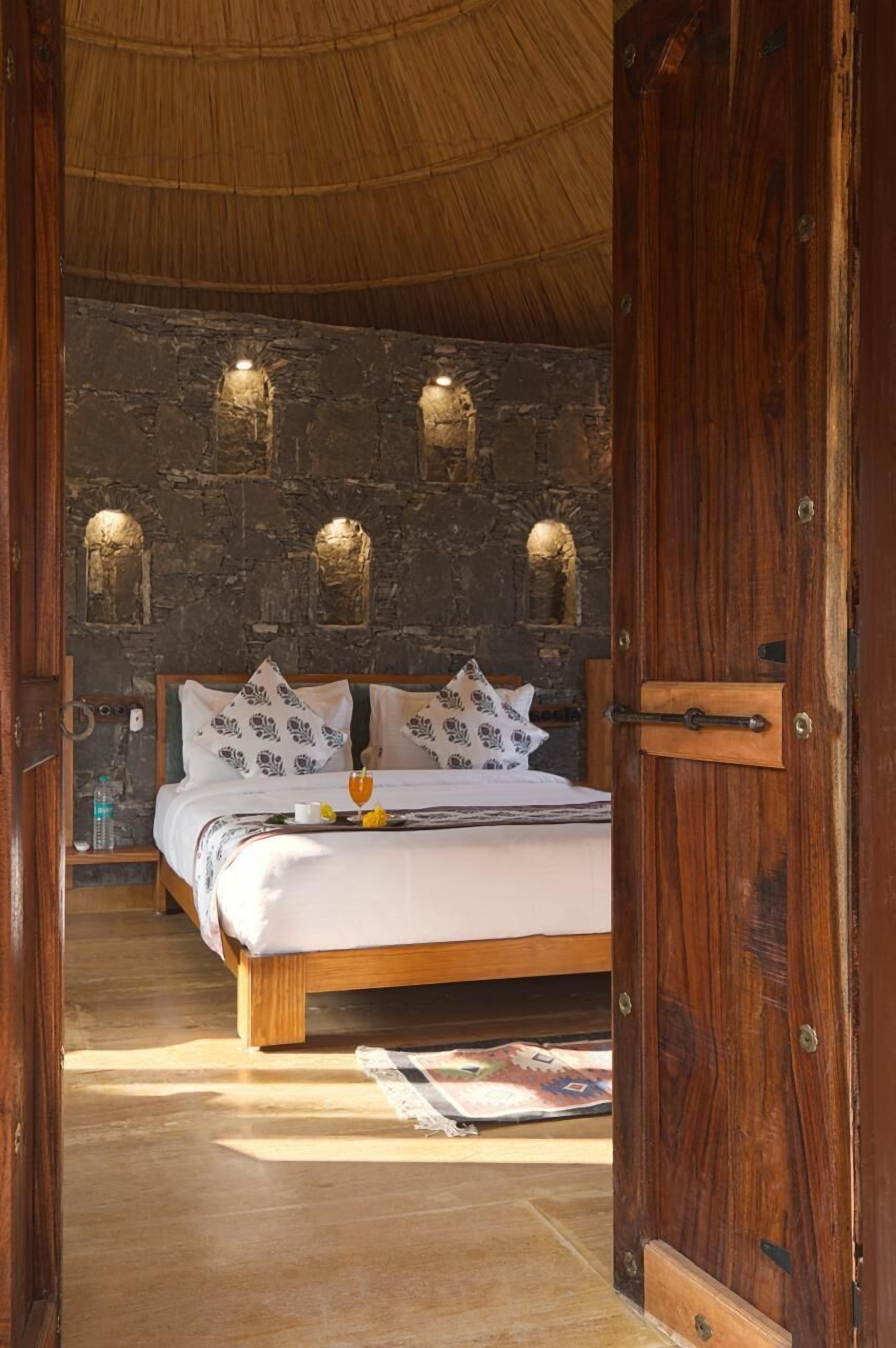 A wooden-framed bed with white bedding and patterned pillows, a stone wall with built-in lighting, and a wooden door.